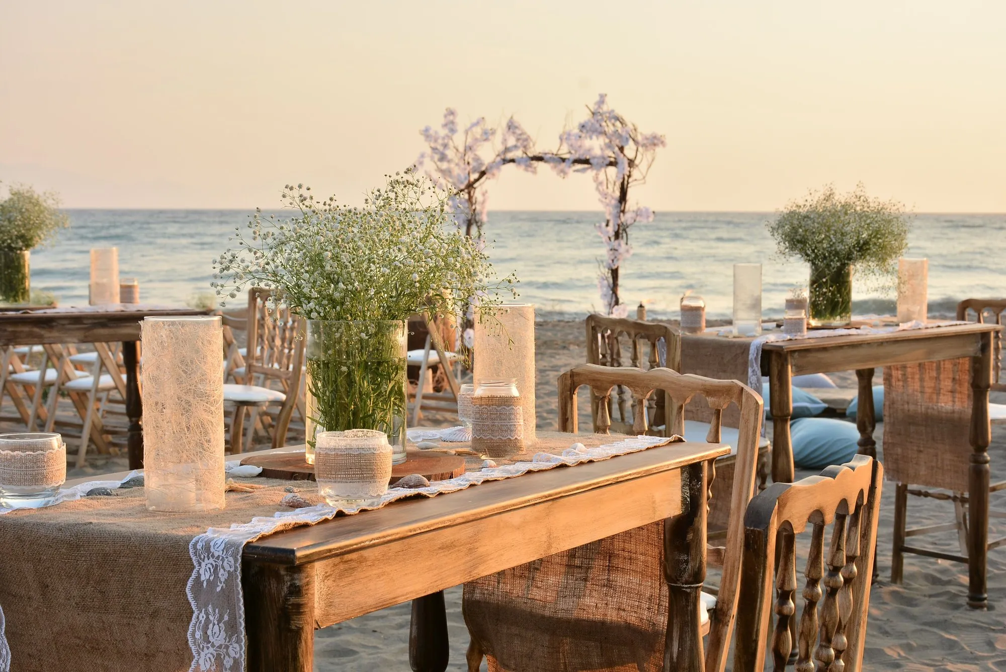 Beach wedding set-up at sunset, with dining tables, ceremony chairs and a flower arch by the sea